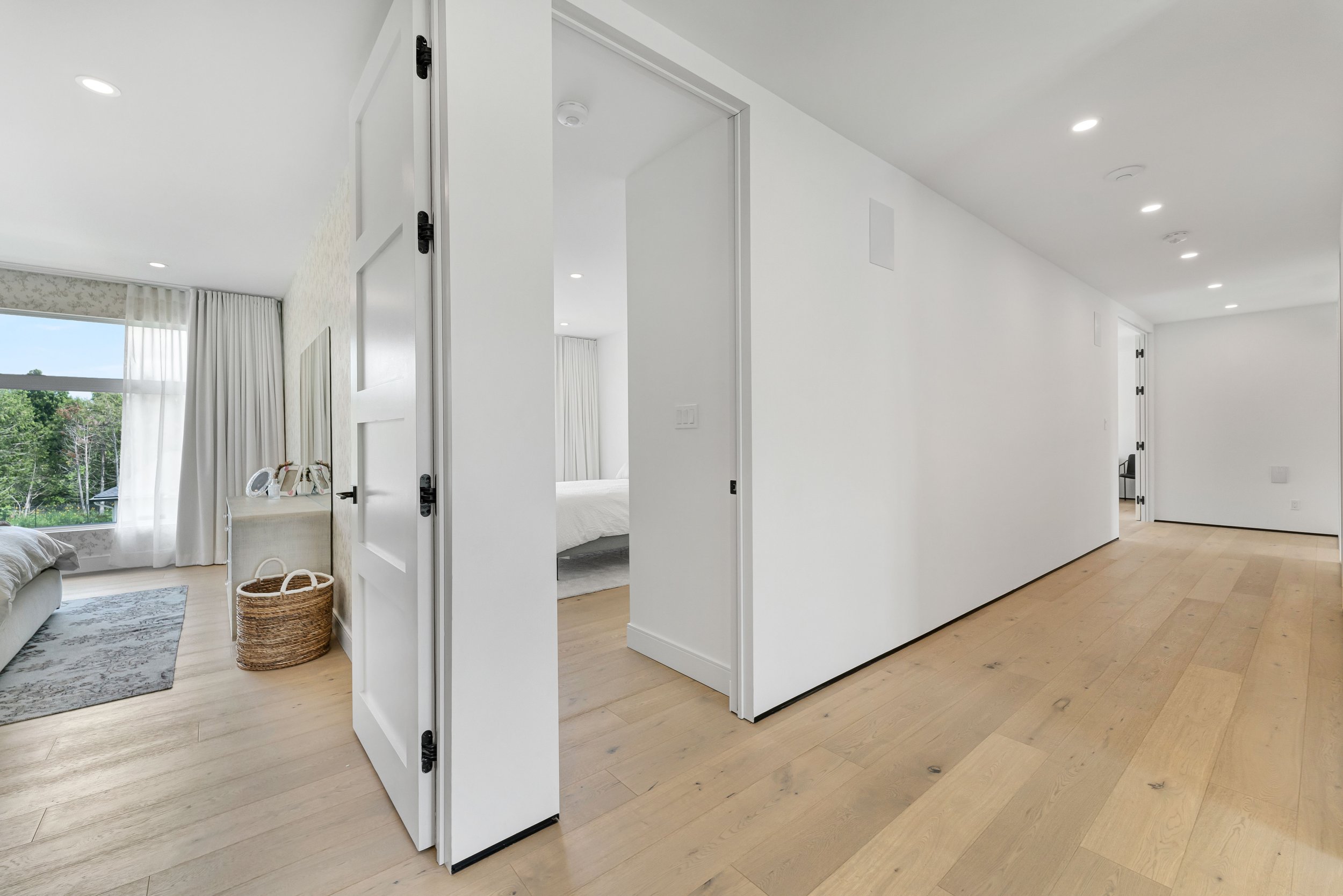 Modern hallway with white walls, wooden flooring, and open doors leading to bedrooms. One bedroom visible with a bed, curtains, mirror, and a wicker basket. Large windows offer a view of trees.