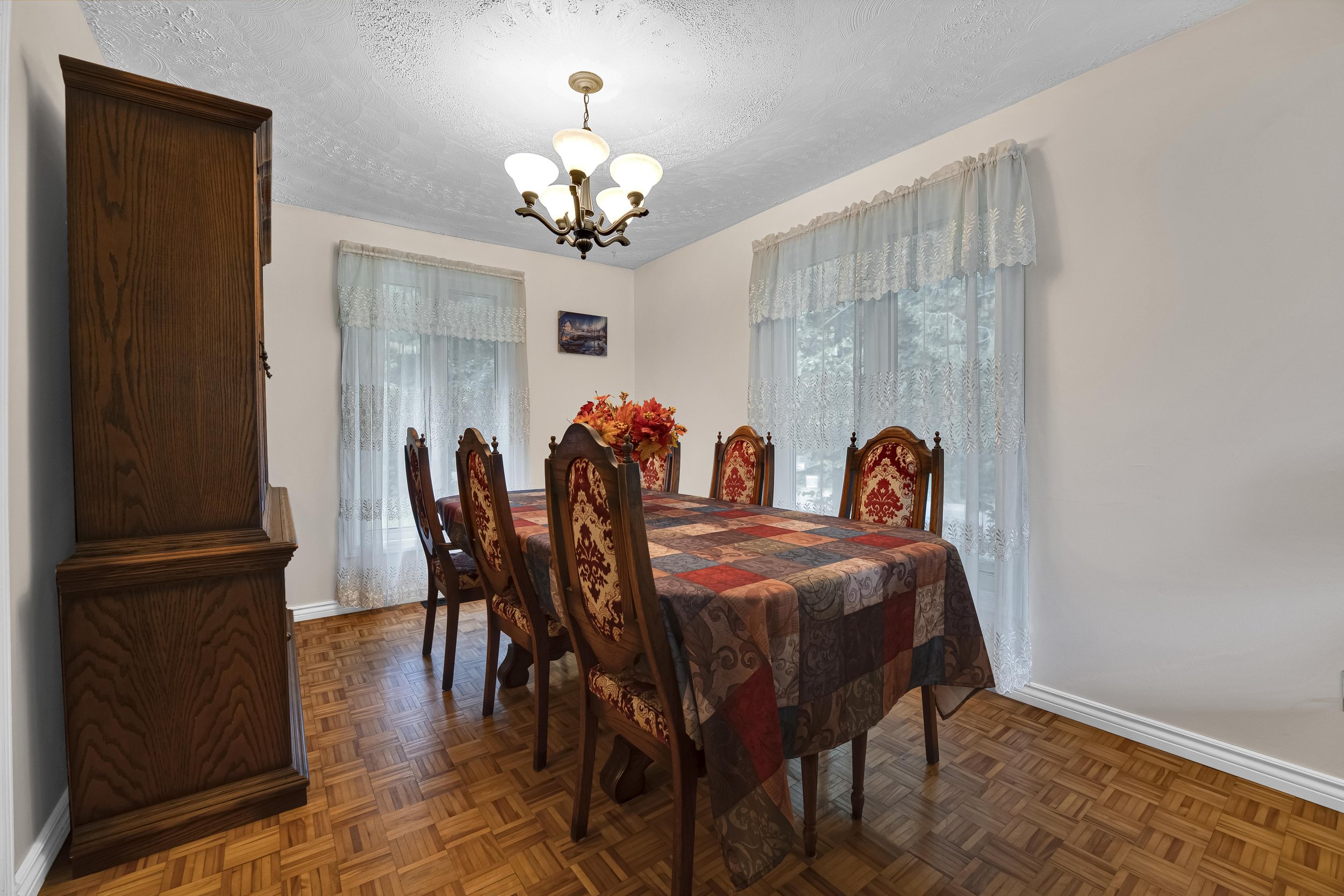 A dining room with a wooden dining table covered with a multicolored tablecloth, six upholstered chairs with a floral pattern, a chandelier hanging from the ceiling, large windows with lace curtains, a wooden cabinet, and a small landscape painting o