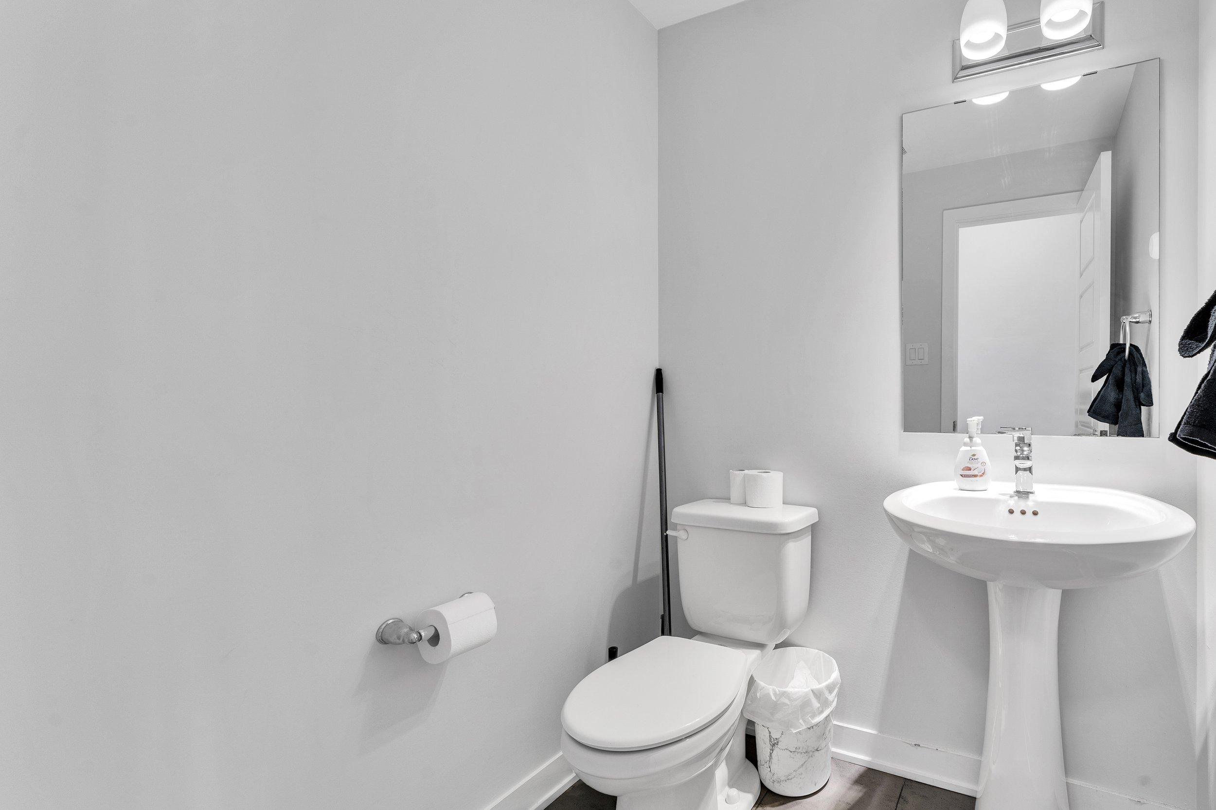 NV3D Real Estate Photography Ottawa  A minimalistic bathroom with a white toilet, a pedestal sink with soap dispenser, a mirror, and a trash can. The walls are painted white and there is a light fixture above the mirror.