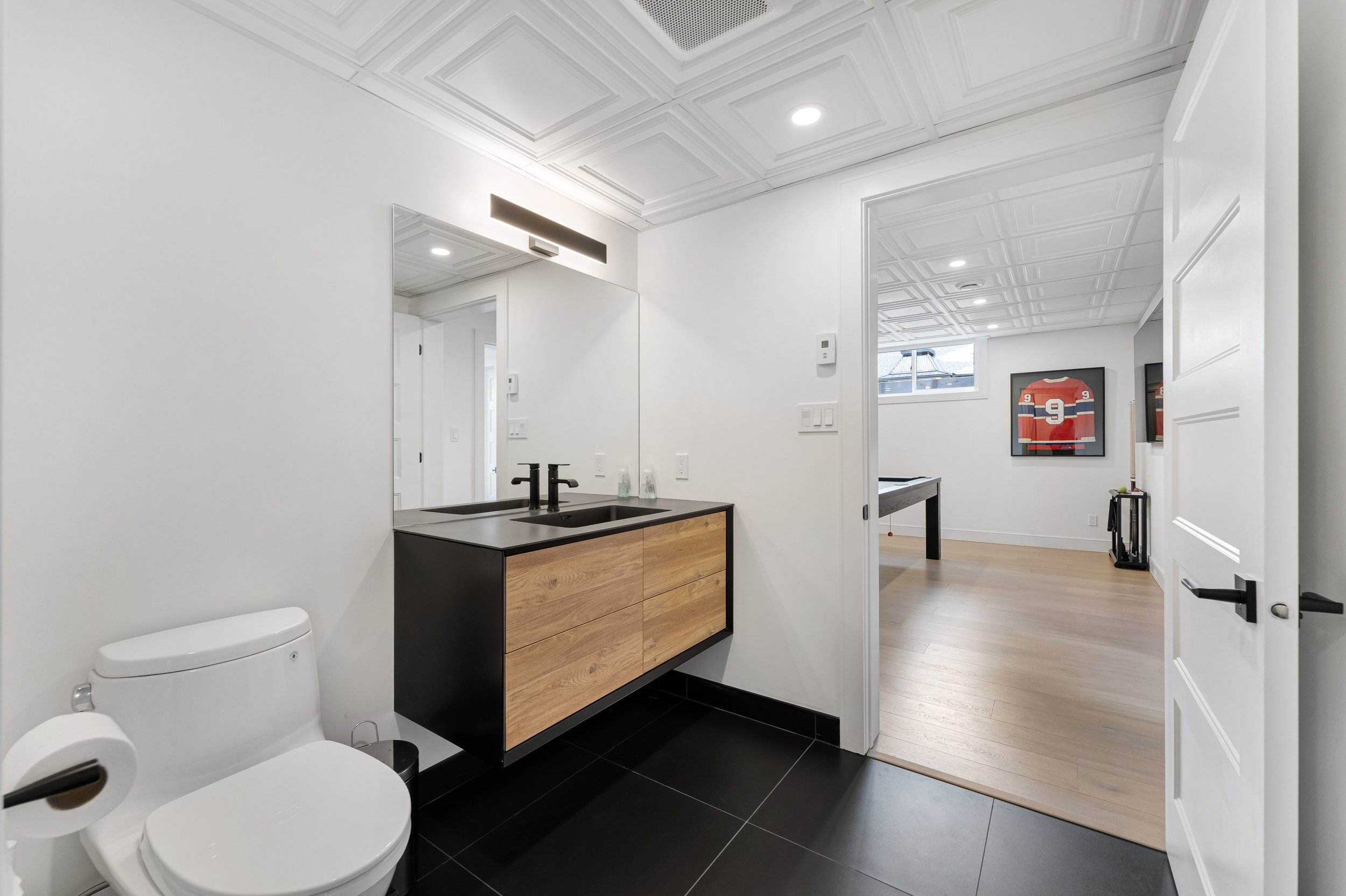 Modern bathroom with black and wood vanity, toilet, and large mirror; adjacent room with framed sports jersey and pool table.