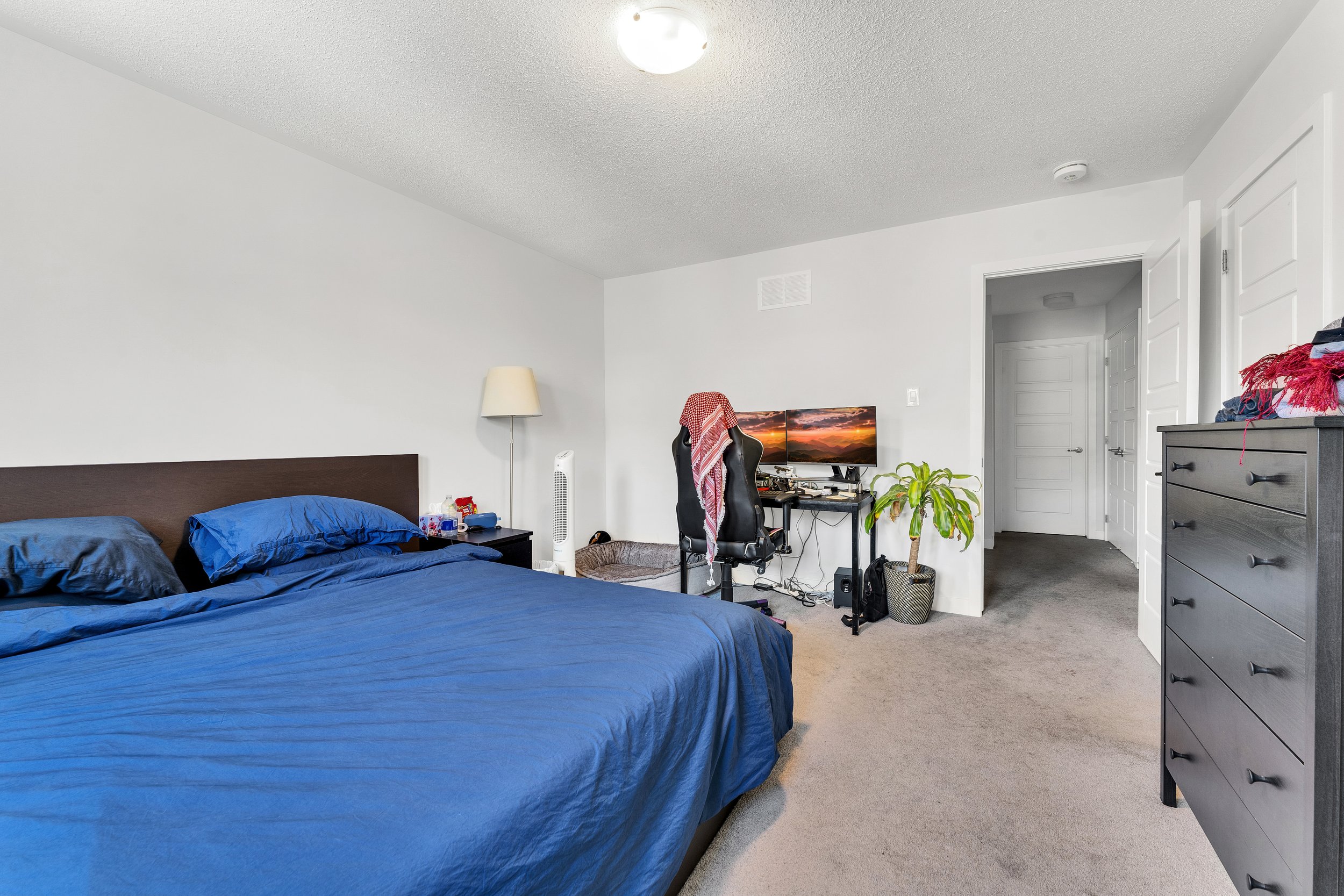 A bedroom with a bed in the left corner covered with a blue comforter and matching pillows. Next to the bed is a small nightstand with various items and a lamp. In the corner, there is a tall fan. On the right side, there is a black dresser with fold