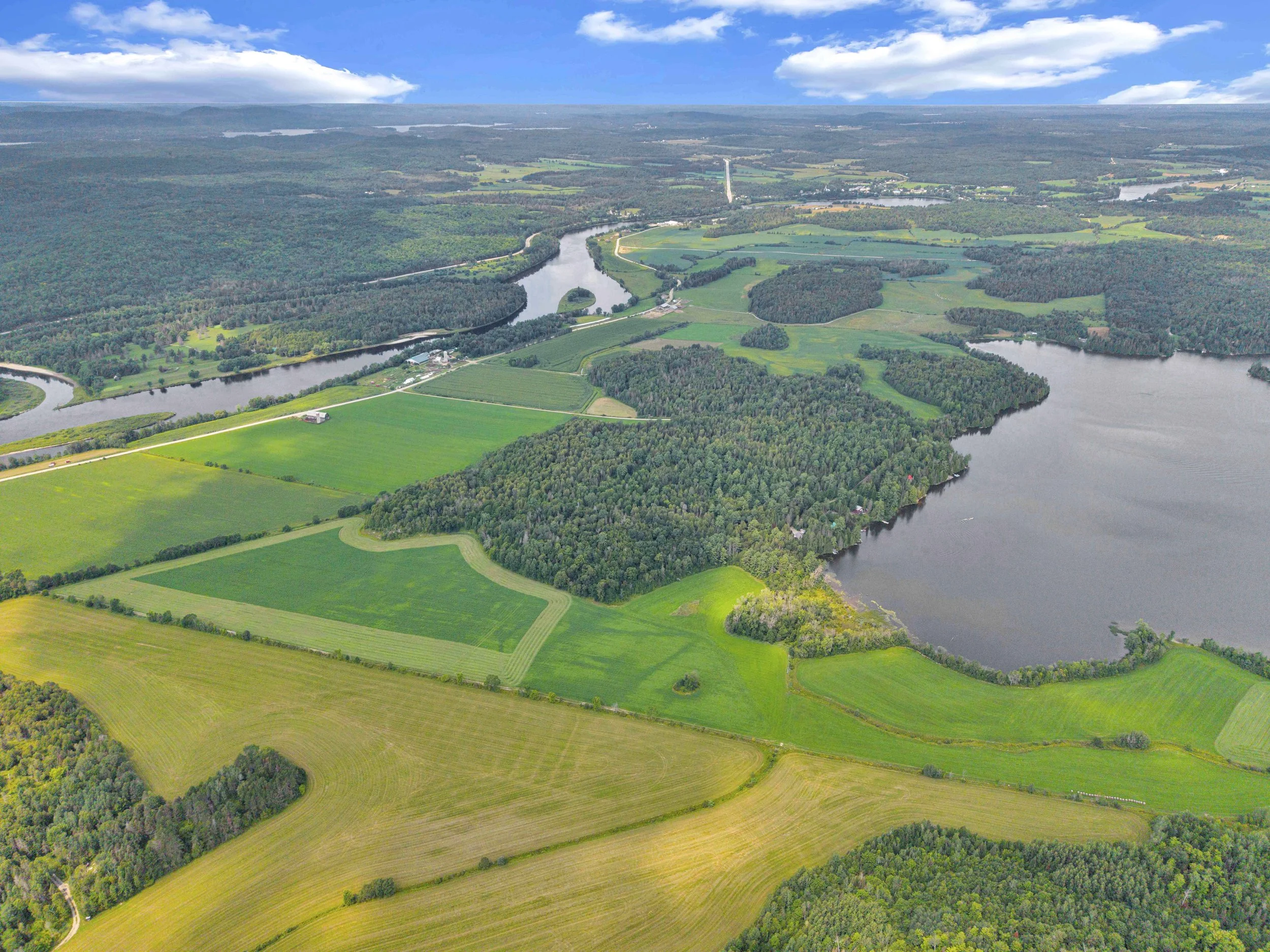 Aerial view of a landscape featuring multiple green fields, dense forests, and winding rivers under a partly cloudy blue sky.