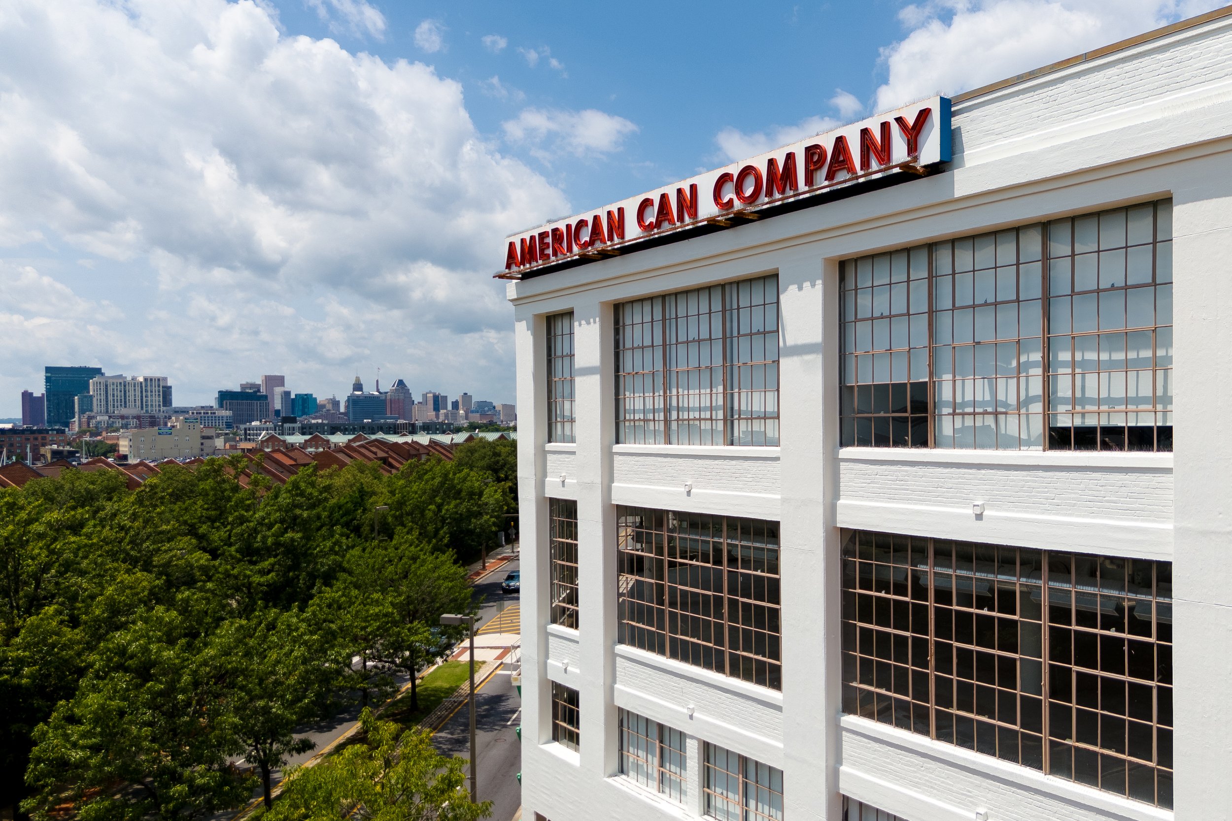 An industrial-style office or creative workspace with large windows, scattered furniture, and various clutter including boxes, a ladder, and equipment.