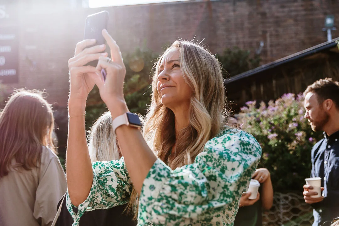 Woman with long blonde hair smiling and taking a photo with her smartphone outdoors during daylight, with sunlight flare and other people in the background holding coffee cups.