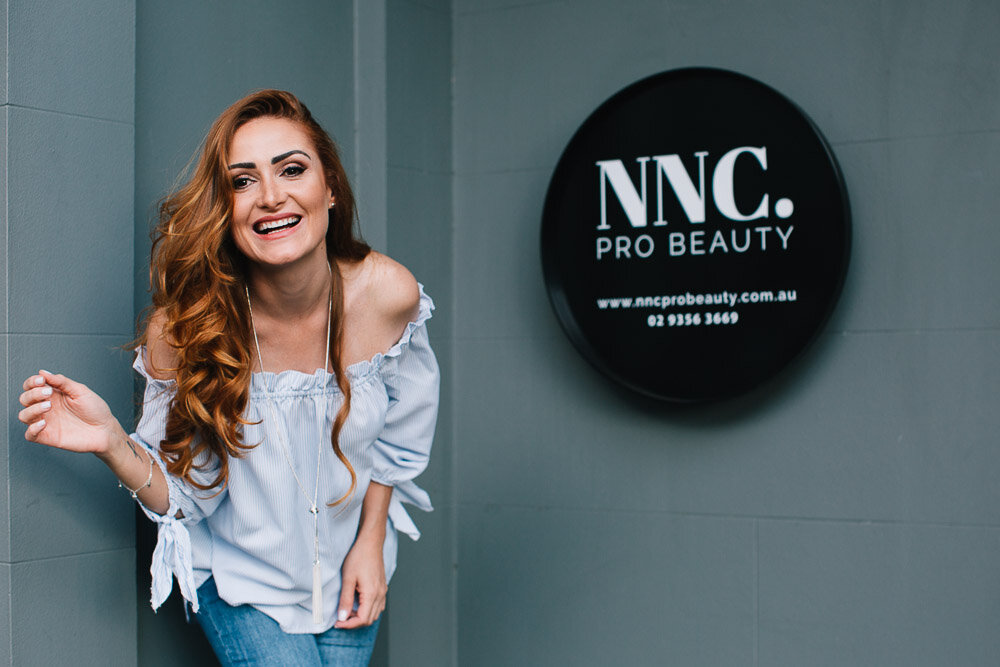 A woman with long red hair smiling and leaning against a wall with a sign that reads 'NNC. PRO BEAUTY' and a website URL in front of a gray wall.