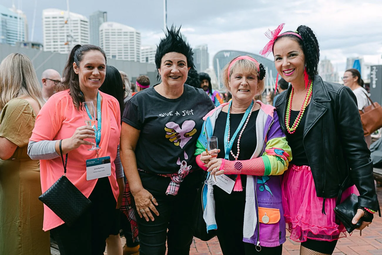 A group of four women smiling at an outdoor event, holding glasses of wine, with a city skyline in the background.