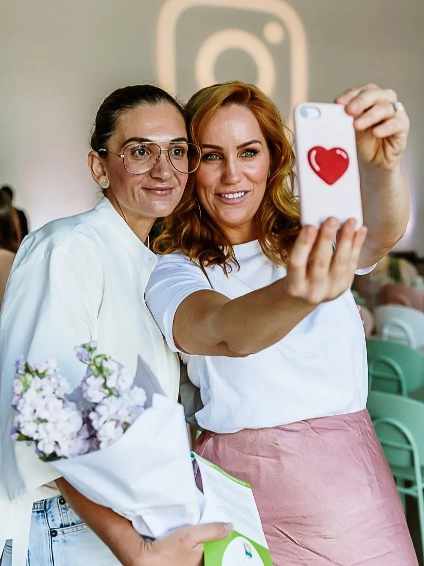 Two women taking a selfie together, one holding a phone with a heart on the case, in a well-lit indoor space with a large Instagram logo in the background.