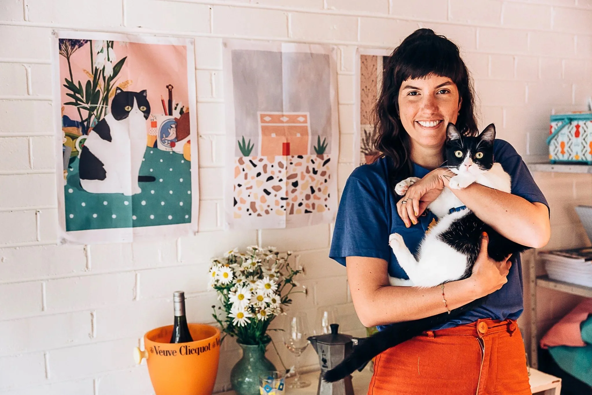 A woman with dark hair and freckles smiling while holding a black and white cat in her arms. Behind her are colorful prints of cats on a white brick wall, a bouquet of daisies, and a table with a bottle of Veuve Clicquot champagne.