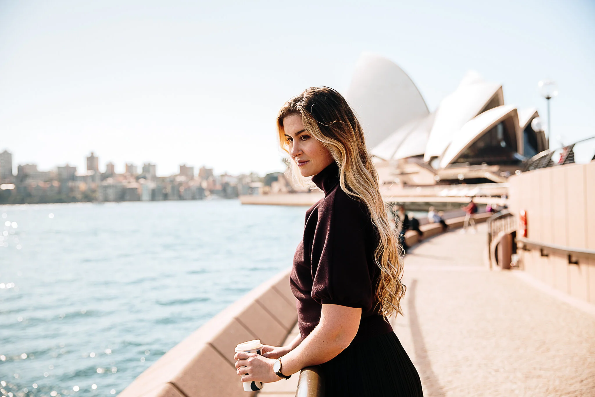 A woman with long hair looking down while holding a coffee cup and standing by the water with a city skyline and the Sydney Opera House in the background.