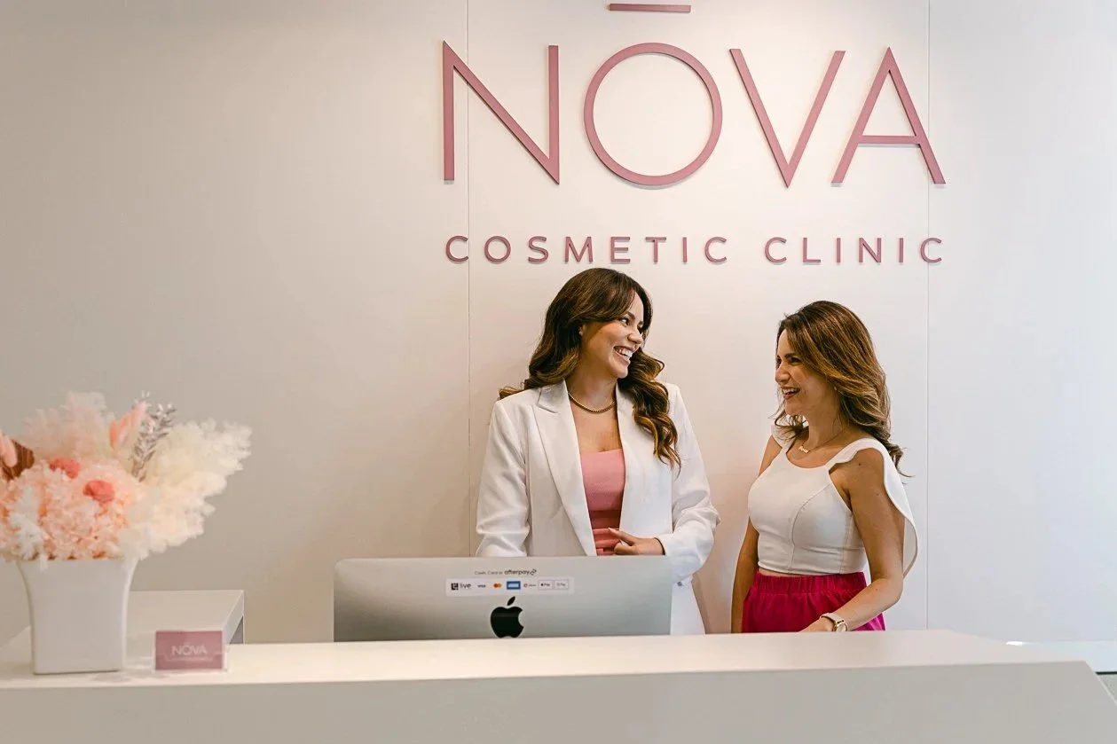 Two women smiling and talking at the reception desk of NOVA Cosmetic Clinic, with a computer, a flower arrangement, and a sign on the desk.