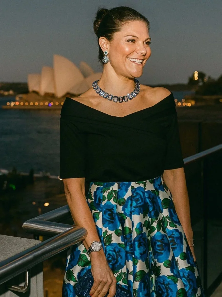 A woman in an off-the-shoulder black top and a blue floral skirt standing on a balcony at dusk with the Sydney Opera House in the background, wearing jewelry and holding a blue clutch purse.
