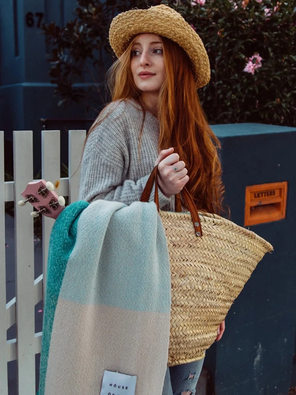 Young woman with long red hair wearing a straw hat and gray sweater, holding a large straw tote bag and a blanket, standing near a white fence and a blue wall with pink flowers in the background.