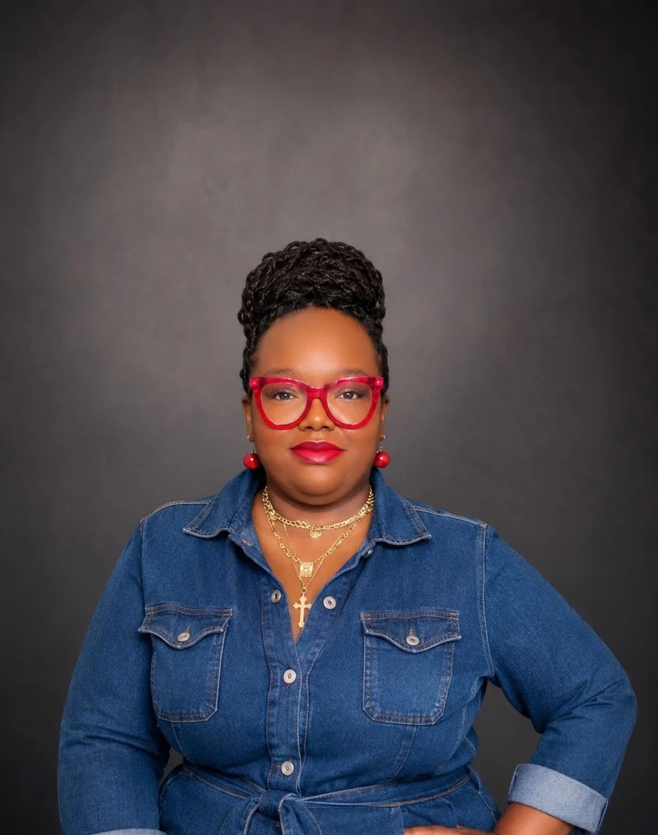 A woman with dark brown hair styled in twists, wearing red glasses, red earrings, gold necklaces, and a denim shirt, stands against a gray background.