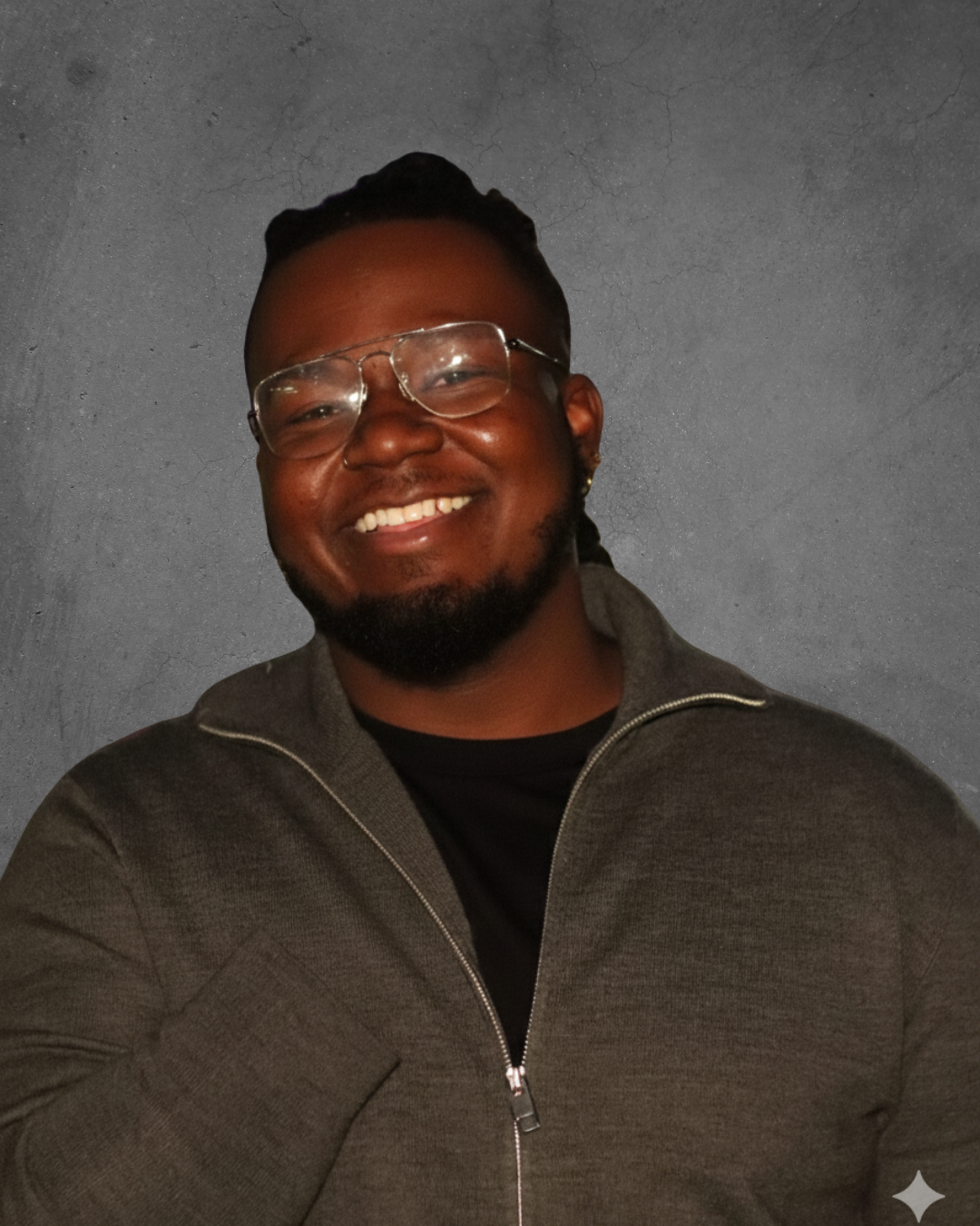 A smiling man with glasses and a beard wearing a dark zip-up jacket against a textured gray background.