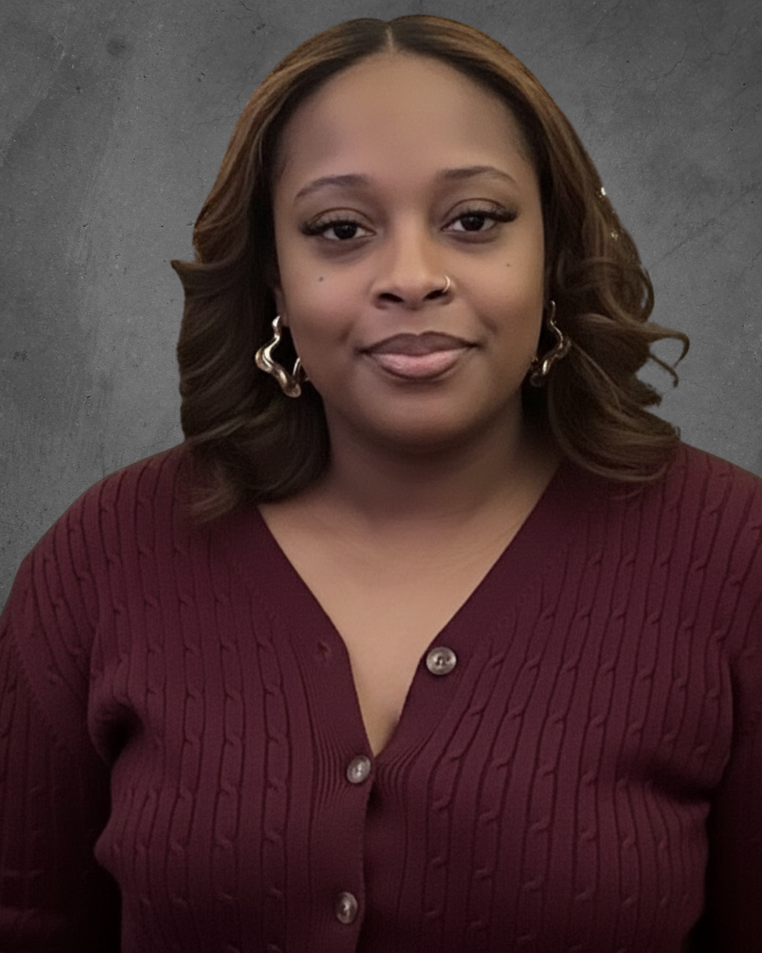 Portrait of a woman with brown hair, wearing a maroon cardigan with buttons and hoop earrings, standing against a gray background.