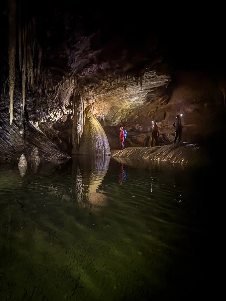 A trio of cavers pause at the water's edge as a floodlit flowstone curtain doubles itself in the still, emerald pool below. Croesus Cave,  Mole Creek Karst National Park, Tasmania.