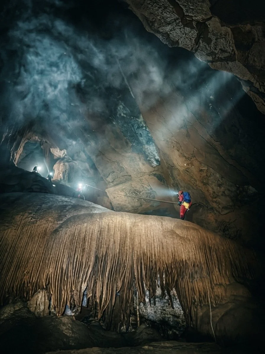 Cavers traverse a curtain of drapery formations, headlamp beams slicing through drifting mist as shafts of light break through the ceiling above. Kubla Khan Cave, Mole Creek Karst National Park, Tasmania.