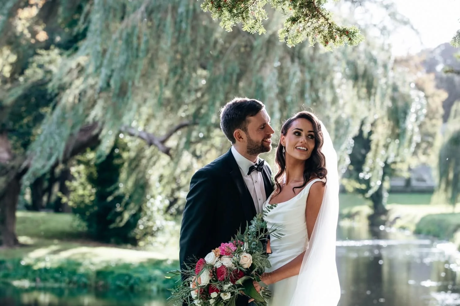 Bride and groom standing outdoors near a pond with trees in the background, the bride holding a bouquet of flowers and smiling, the groom dressed in a tuxedo, enjoying a wedding moment.