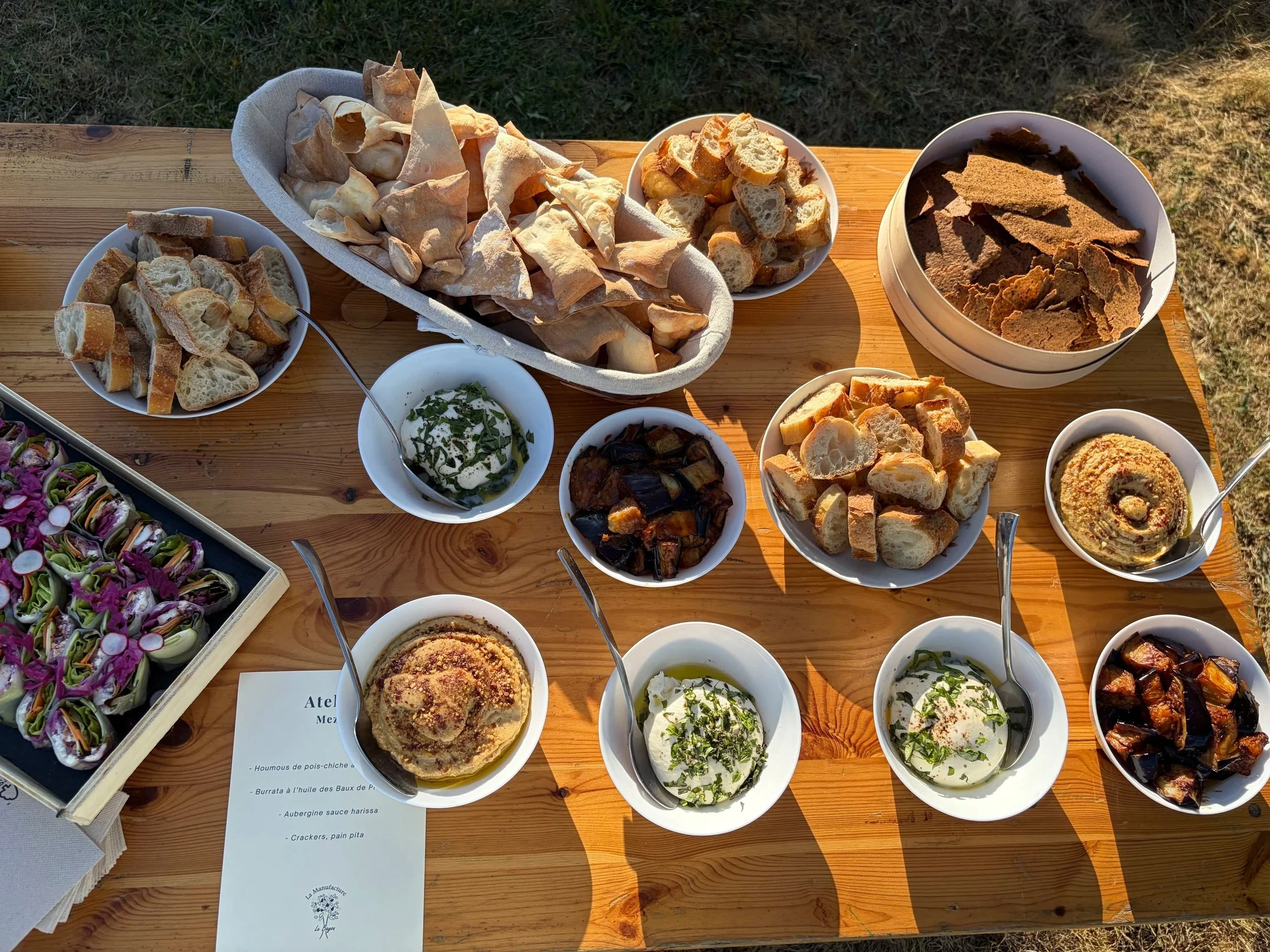 Assortiment de pain, tartines, dips, et snacks sur une table en bois, en plein air.