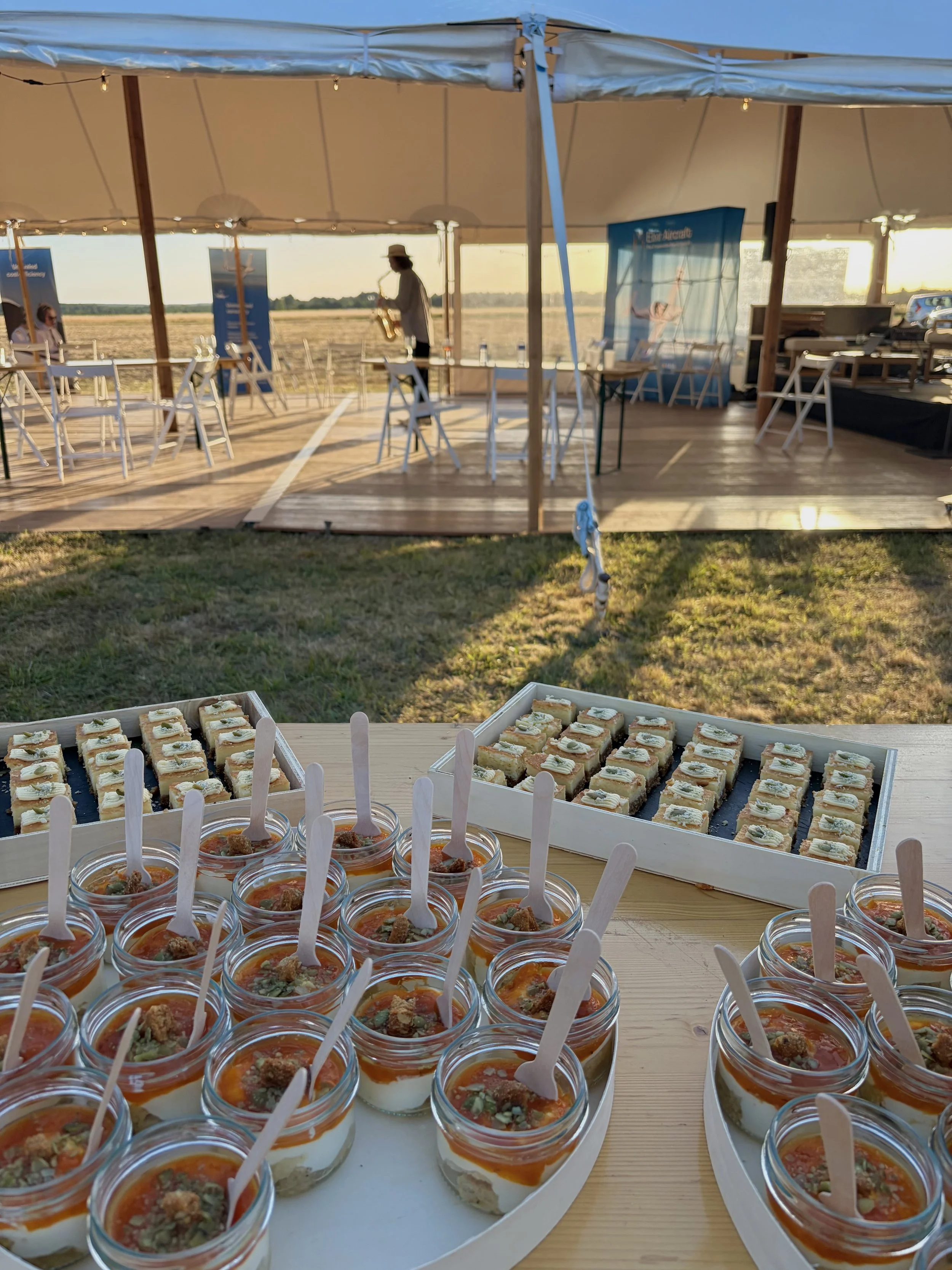 Apéritifs et amuse-bouches servis dans de petits verres et sur des plateaux, avec un fond d'une tente en plein air au coucher de soleil, un groupe de personnes joue de la musique, un saxophoniste en silhouette, sur un terrain herbeux