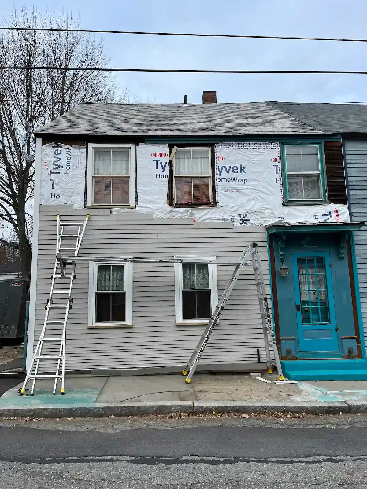 The upper part of a house undergoing renovation with exposed Tyvek HomeWrap, two new windows, and two ladders leaning against the house. The lower part of the house has gray siding and two existing windows, with a new blue front door and small porch.