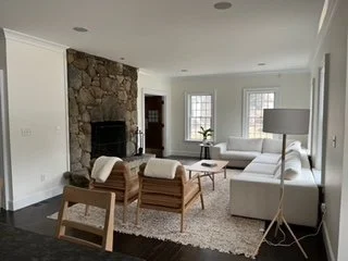 Hampton Swedish inspired living room with a stone fireplace, white sectional sofa, wooden chairs, complemented with white cushions, white rug, floor lamp, and large windows.