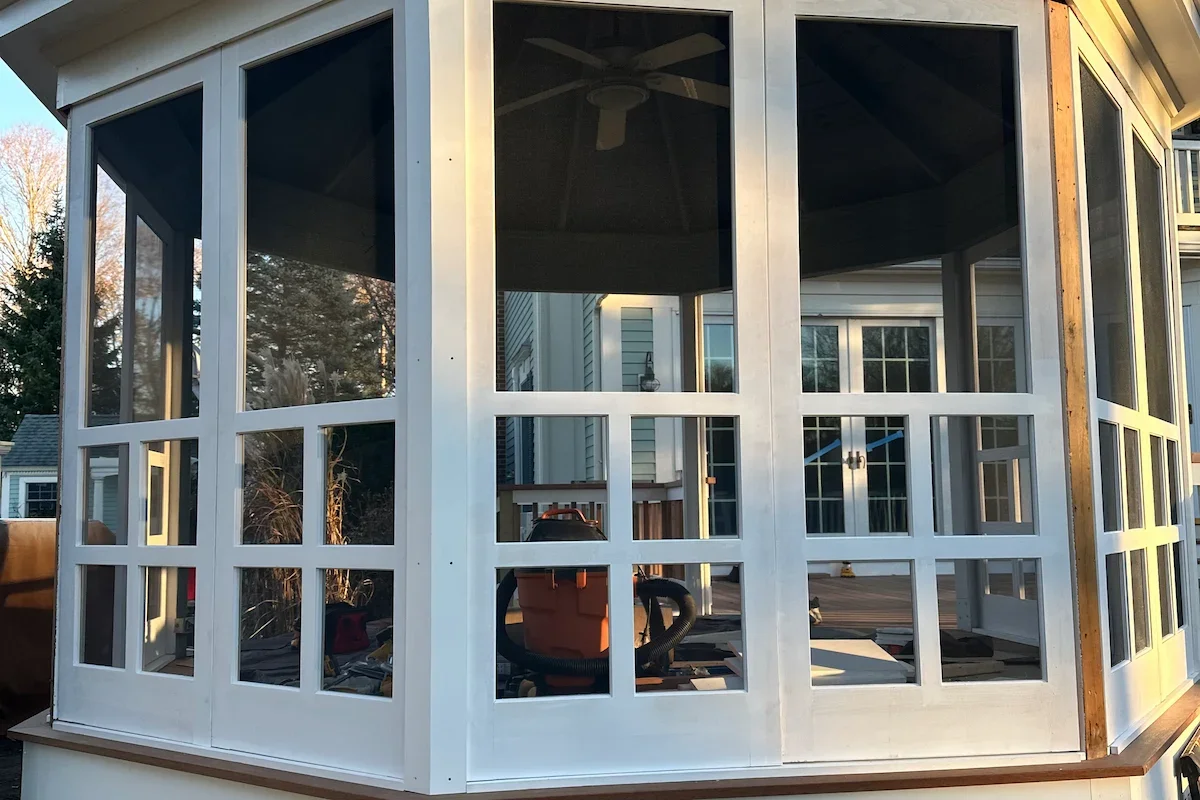 A screened porch or sunroom under construction attached to a house, with white framing and large glass windows, and construction tools and equipment inside.
