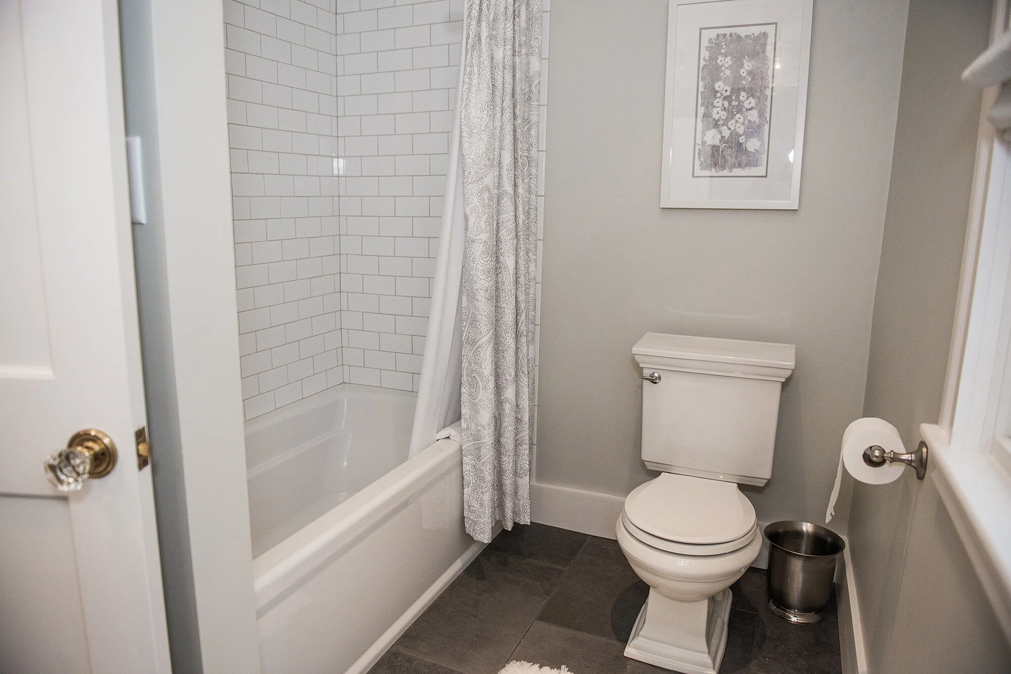 A bathroom in west Newbury  with a white bathtub and tiled shower area, a white toilet, a framed floral artwork on the wall, a silver trash can, and a toilet paper roll on a holder.