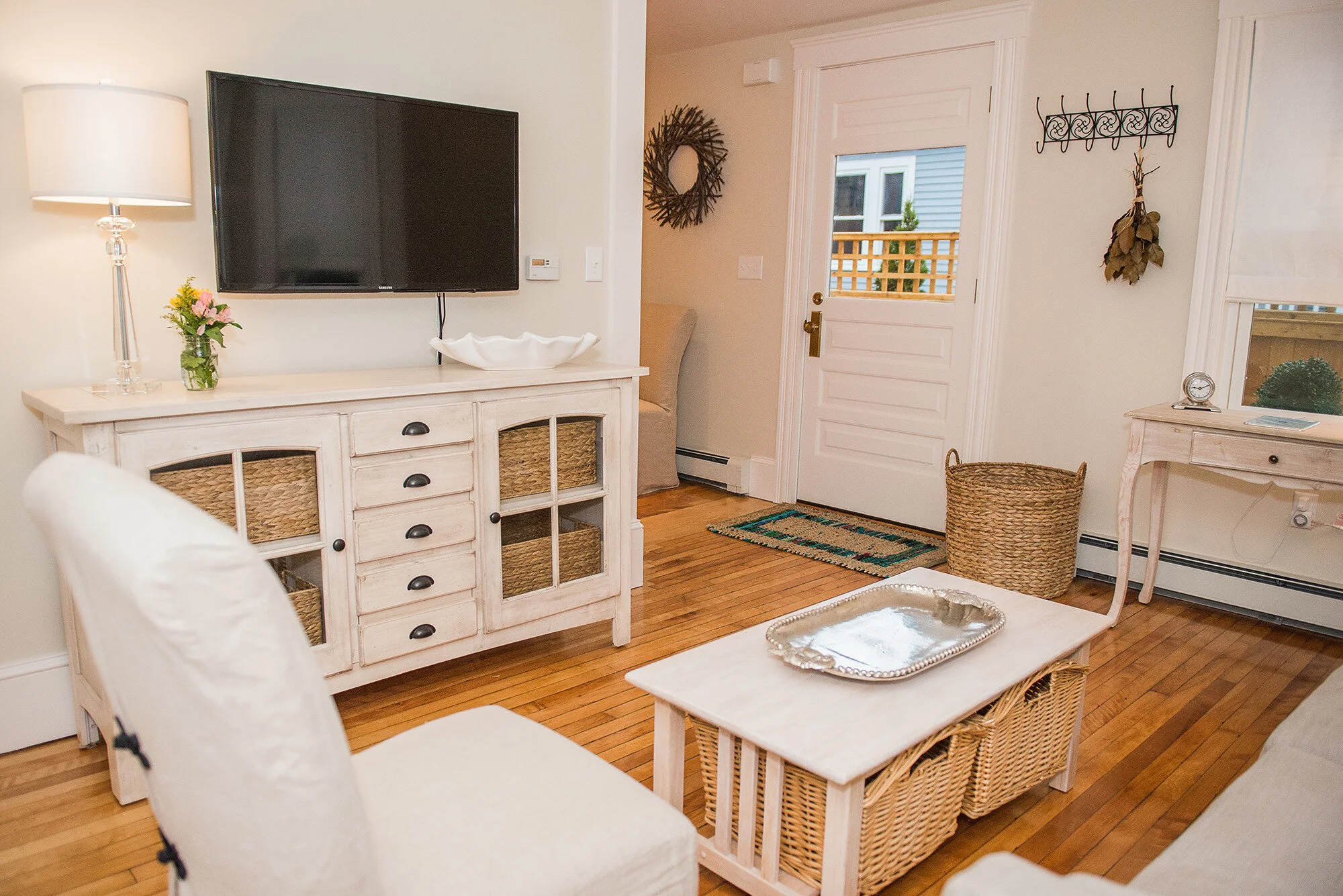 Living room in downtown Newburyport with a white sideboard, a mounted TV, a white table with a silver tray, and wicker baskets, with a door leading outside.