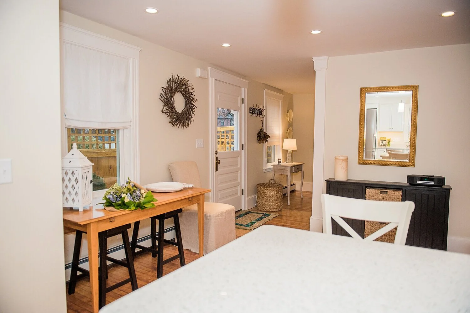 Newbury Interior of a cozy home with a wooden dining table, floral centerpiece, white candle lantern, beige chair, black sideboard with a mirror, small table with lamp, and decorative wall accents in a well-lit room.