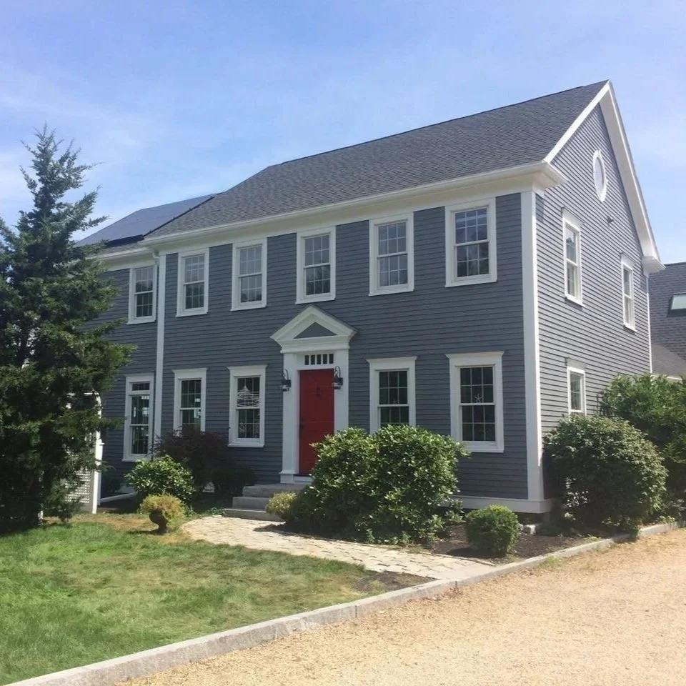 A two-story gray shore front  Newburyport house with white trim and a red front door, surrounded by green bushes and trees, with a gravel pathway leading to the entrance.