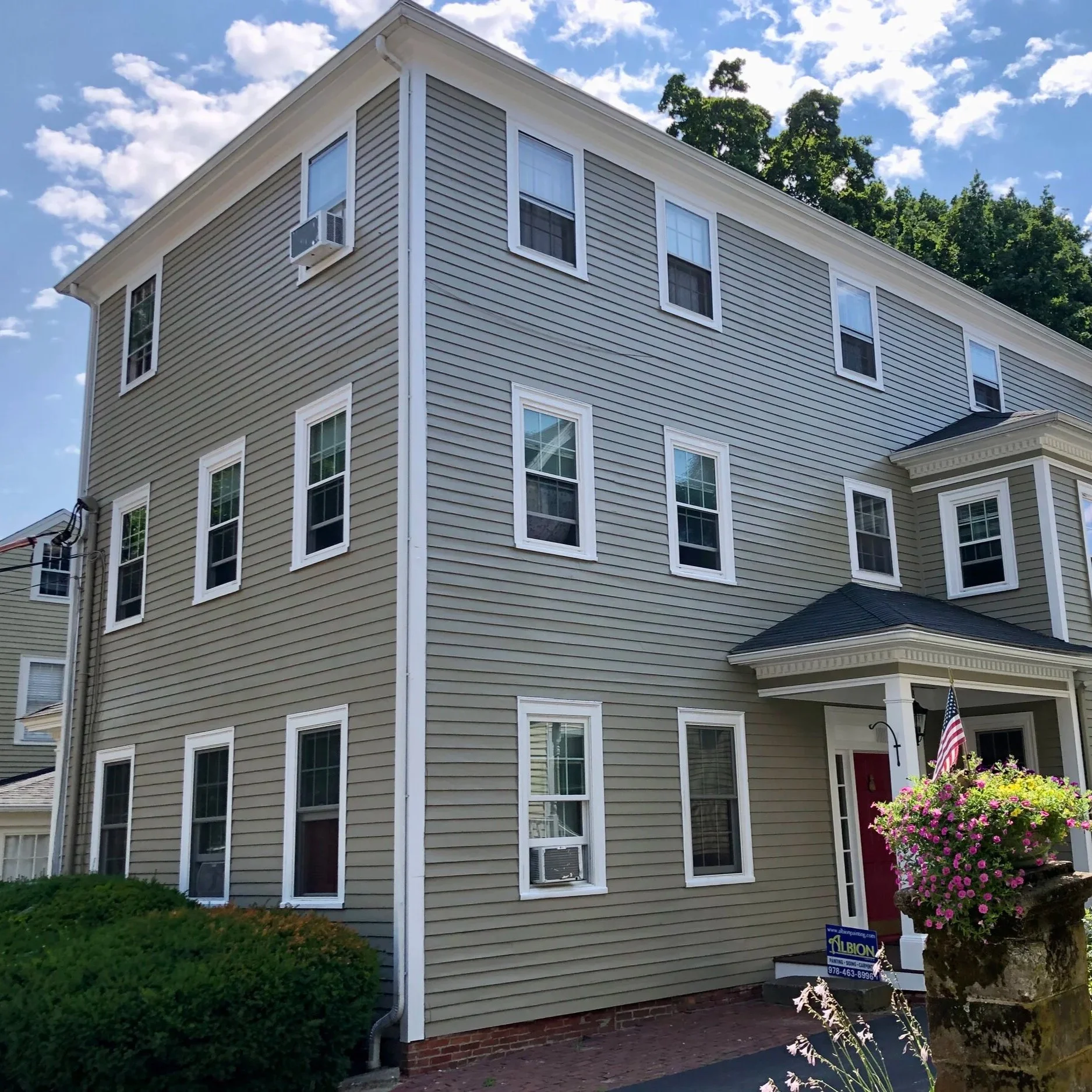 A four-story historic Newburyport house with white trim and multiple windows, surrounded by green trees and a landscaped yard with bushes and flowers. There is a small porch with an American flag and a pink door.