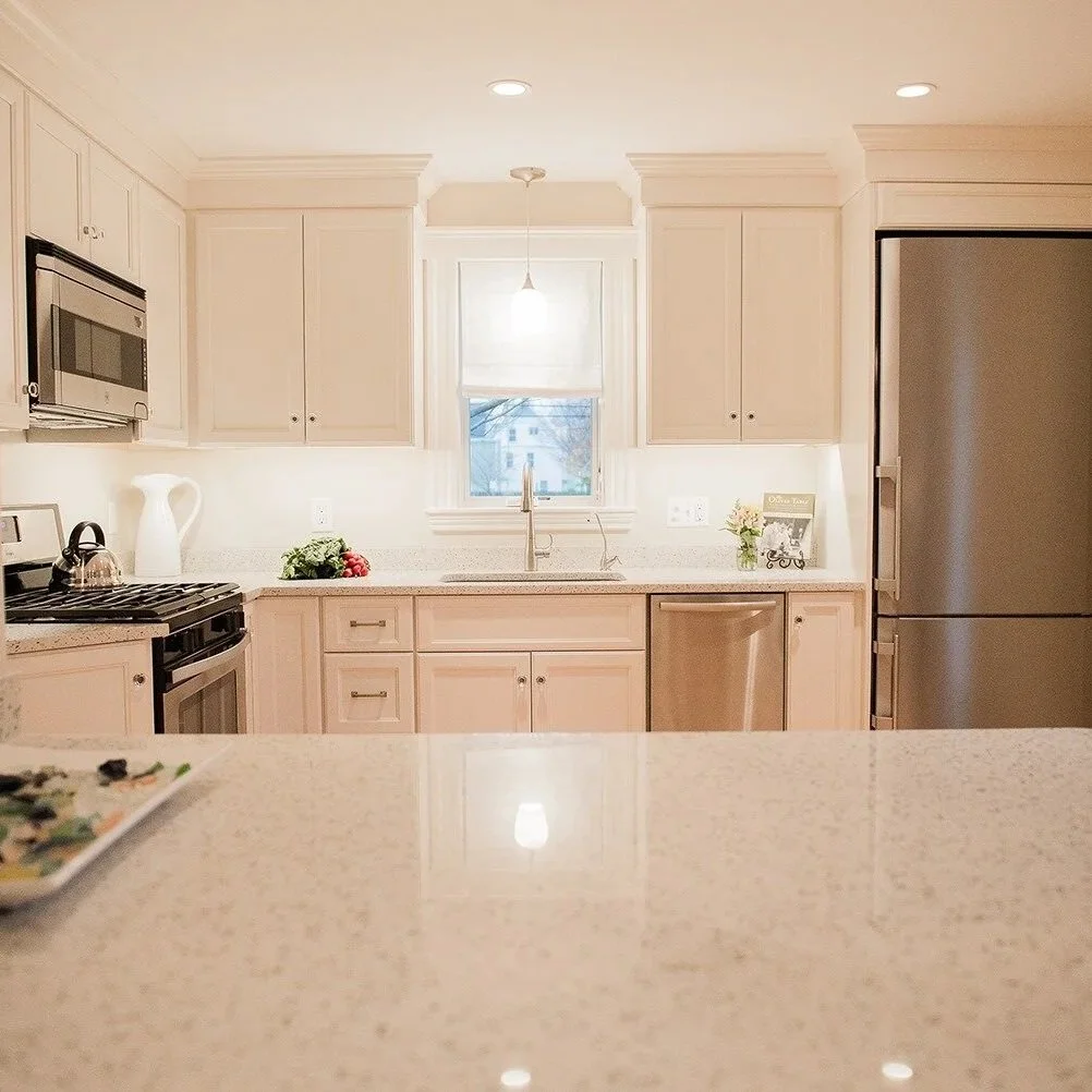 Historic Newburyport  kitchen with stainless steel appliances, including a refrigerator, microwave, and oven, beige cabinets, a window above the sink, small potted plants, a tray with food on the counter, and a hanging light fixture.