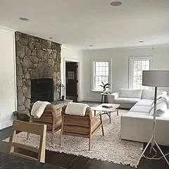 Hampton Swedish inspired living room with a stone fireplace, white sectional sofa, wooden chairs, complemented with white cushions, white rug, floor lamp, and large windows.