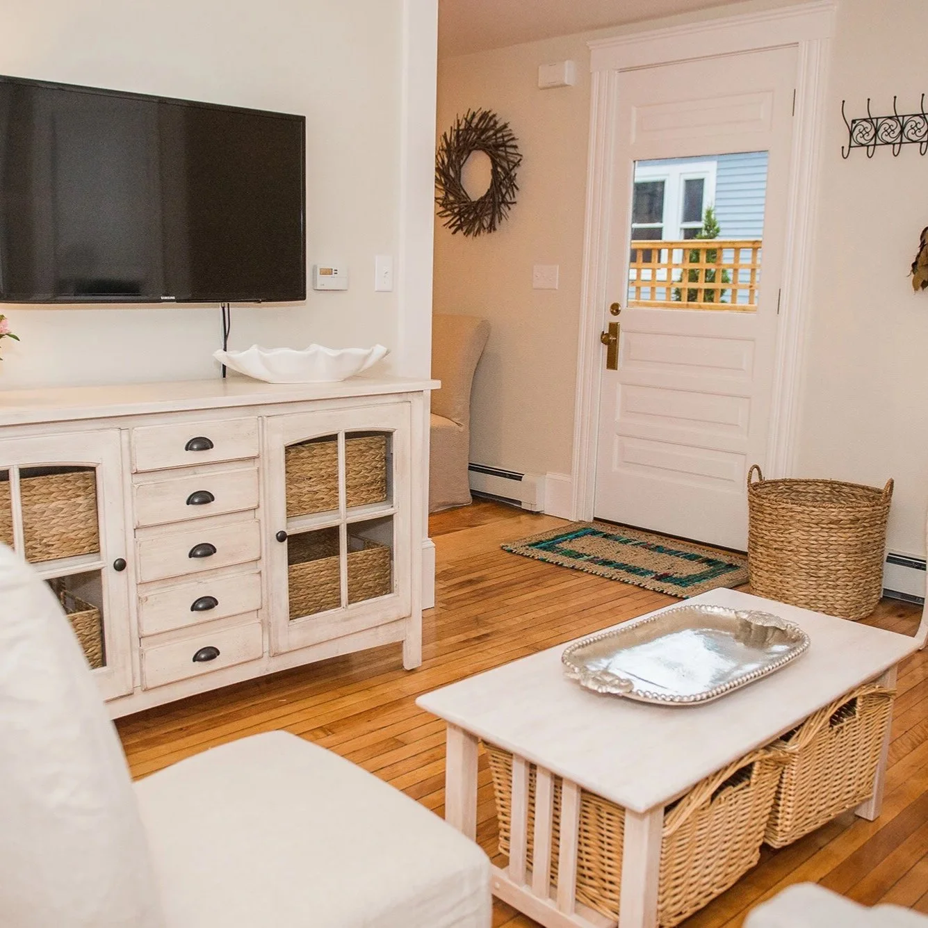 Living room in downtown Newburyport with a white sideboard, a mounted TV, a white table with a silver tray, and wicker baskets, with a door leading outside.