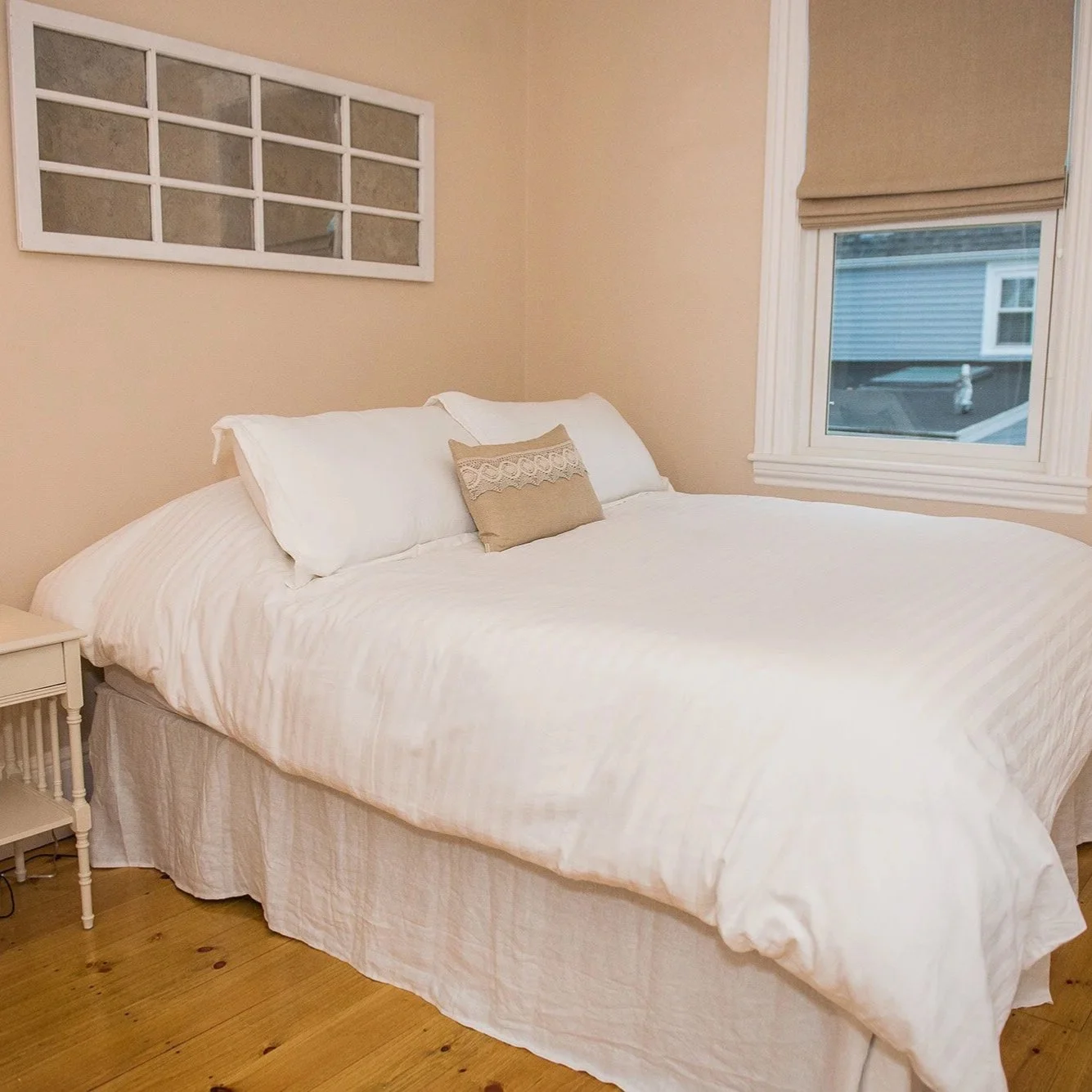 Cosy Newburyport bedroom with a large bed covered in white bedding and pillows, a bedside table with a lamp and a clock, a window with a brown shade, and wooden flooring.