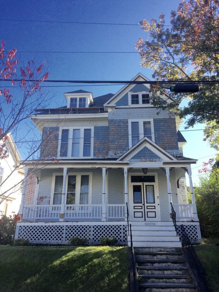 A large VicNewb, three-story house with  blue and white paint, a front porch with white railings, and a staircase leading up to the front door. The house has several windows and a peaked roof, with trees and a clear blue sky in the background.