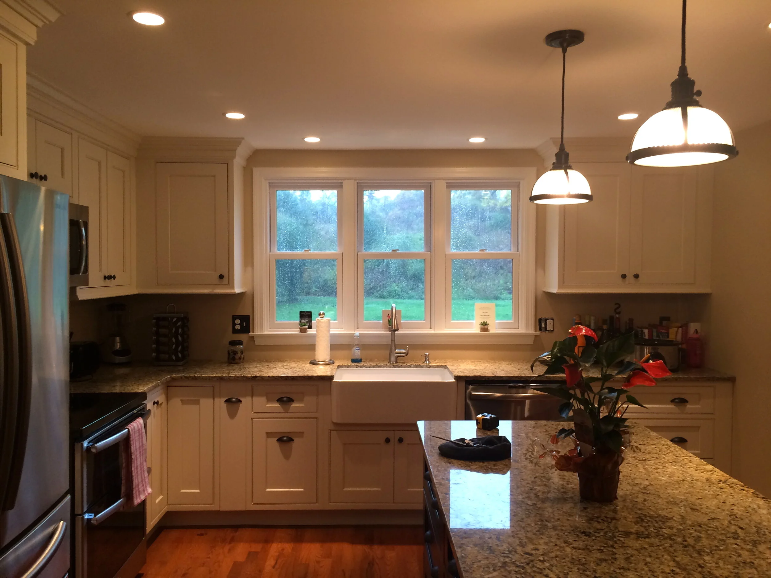 Newbury Kitchen with white cabinetry, granite countertops, a window above the sink looking out to trees, and a kitchen island with a potted plant and a few items on it. Two pendant lights hang over the island.