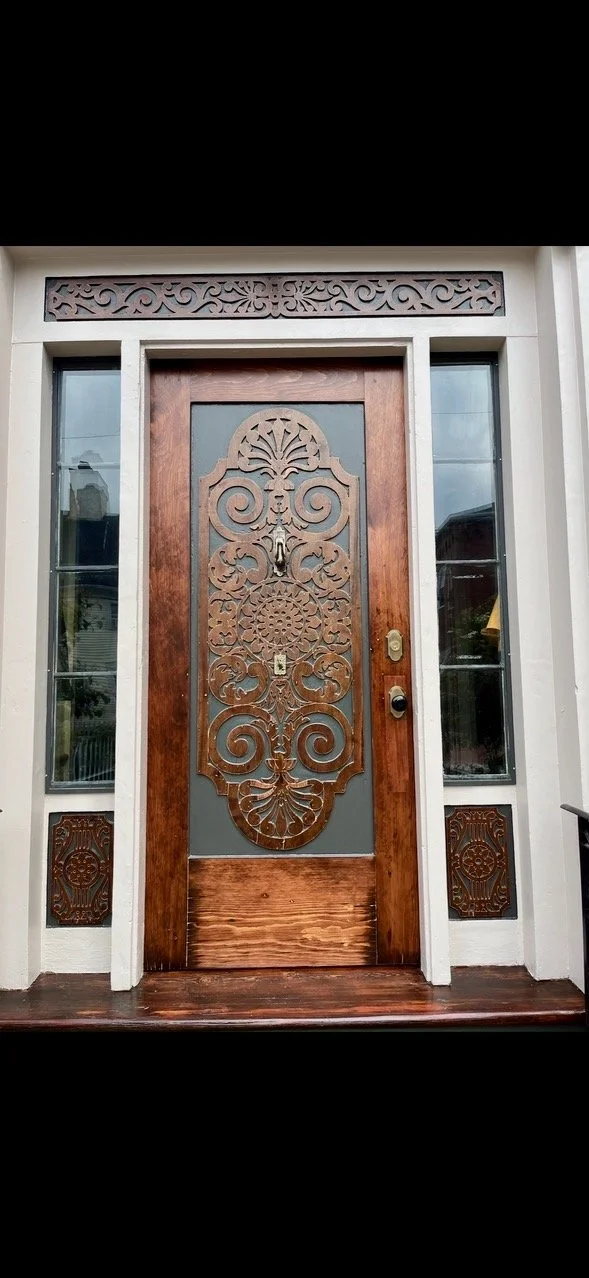 Front door with decorative ironwork and wooden frame, flanked by two narrow glass panels, with ornamental wood carvings above and below.