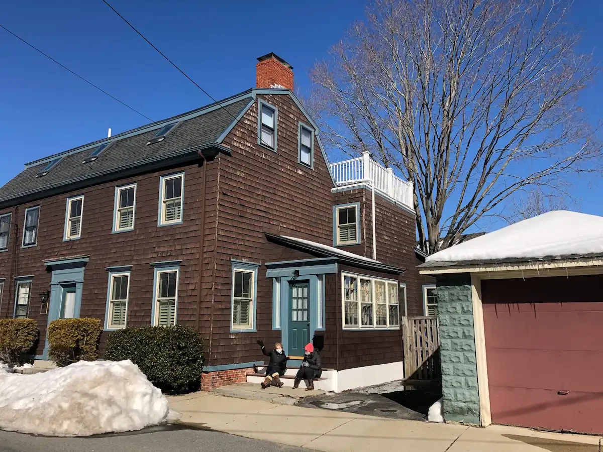 A brown multi-story house with shingle siding, blue window frames, and a brick chimney. Two children wearing winter clothing and masks are sitting on the front steps, with snow on the ground and a large leafless tree in the background.