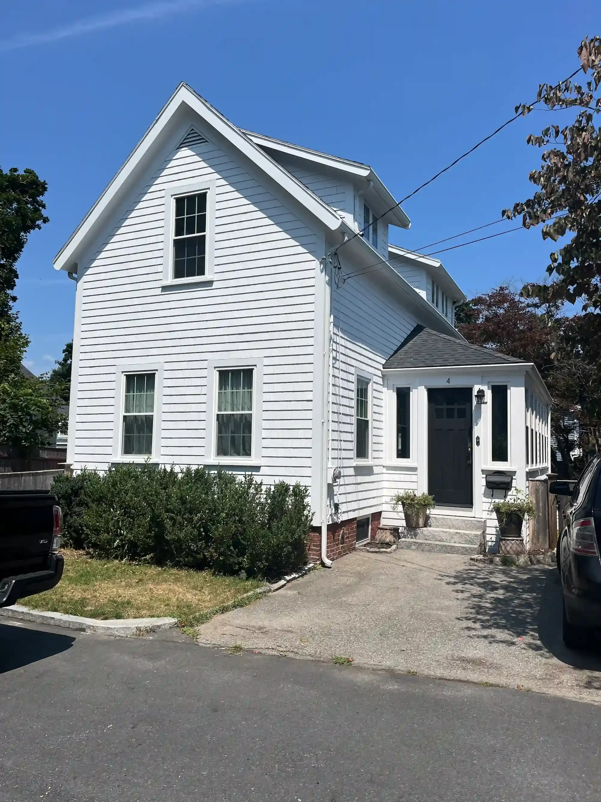 White two-story house with black front door, small porch, and driveways on both sides, surrounded by trees and parked cars, under a clear blue sky.