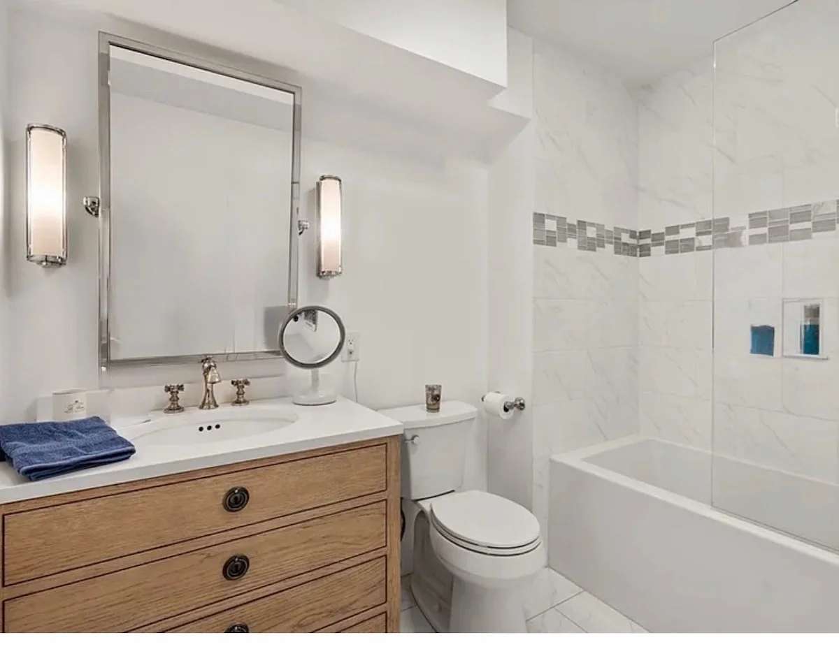 A modern bathroom with a wooden vanity, a mirror, wall-mounted light fixtures, a small round mirror, a toilet, and a bathtub with tiled walls.