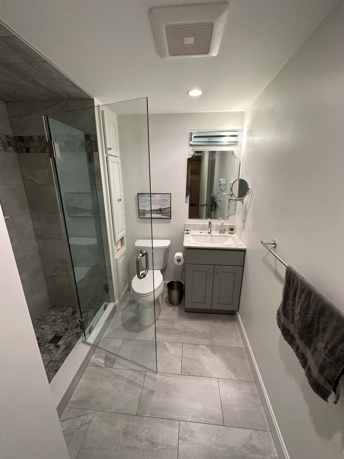 A modern bathroom with a glass-enclosed shower, a toilet, and a vanity with a marble countertop and a large mirror. There is a towel hanging on a rack and a small framed picture on the wall.