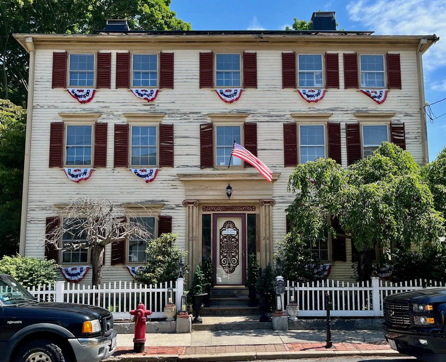 Three-story house decorated with American flags and patriotic bunting for a celebration, with cars parked in front and a white picket fence surrounding the yard.