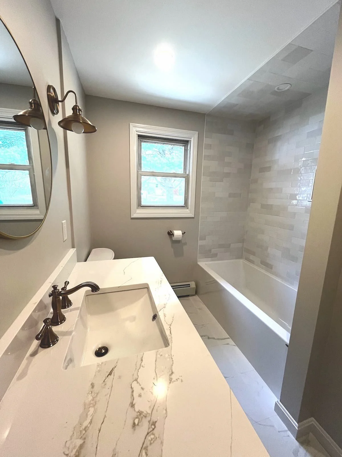Modern bathroom with a white marble vanity and black fixtures, a bathtub with a tiled wall, and two windows. West Newbury Mass