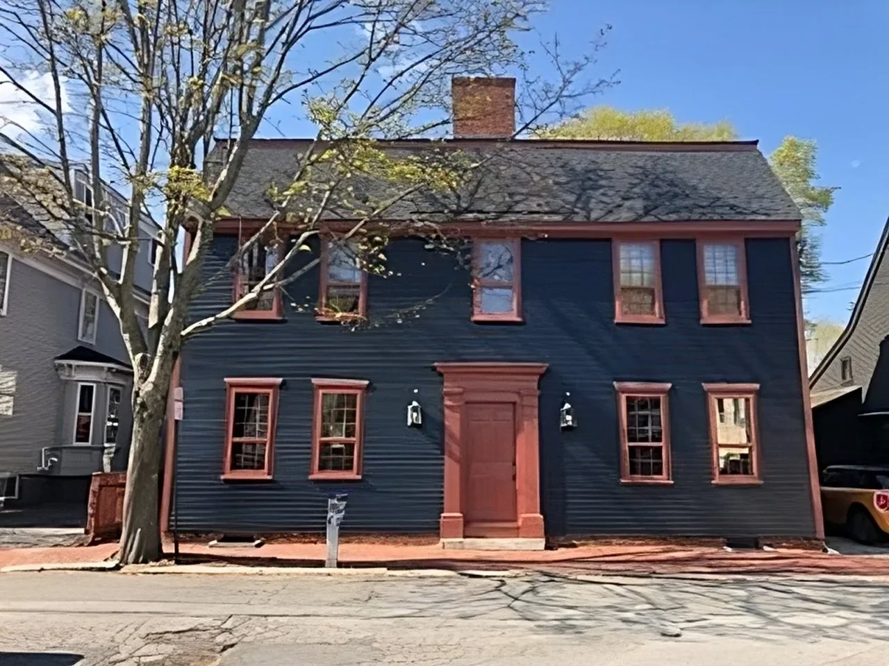 A dark blue wooden house with reddish-brown window frames and door, two black outdoor wall lamps, and a large tree in front. The house has three windows on the upper level and three on the lower level. There is a mailbox in front of the house and a parked vehicle on the right side.