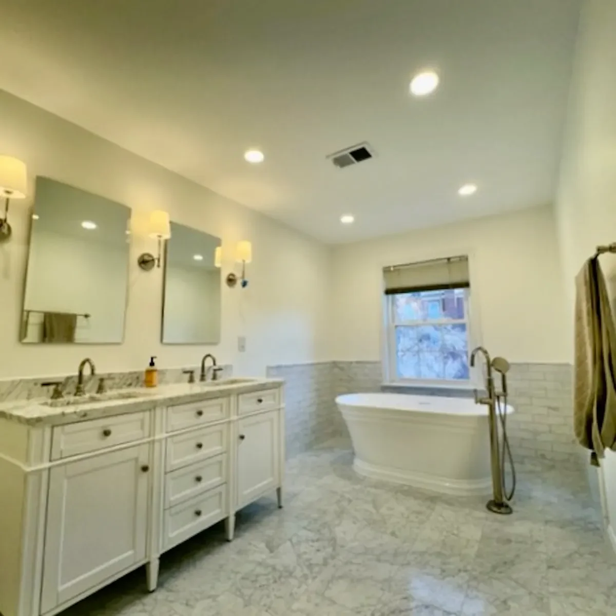 Bright bathroom with double sink vanity, two mirrors, wall-mounted lights, a freestanding bathtub by a window, and a towel rack with towels.