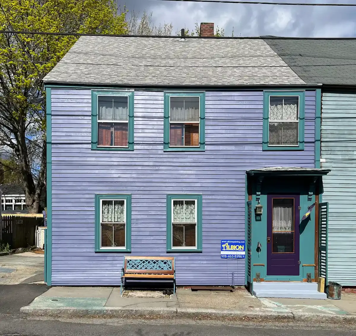 A two-story house painted in pastel purple with green trim around the windows and door. The house has four windows, two on each floor, with lace curtains. There is a small front porch with a purple door and a small bench in front of the house. A sign