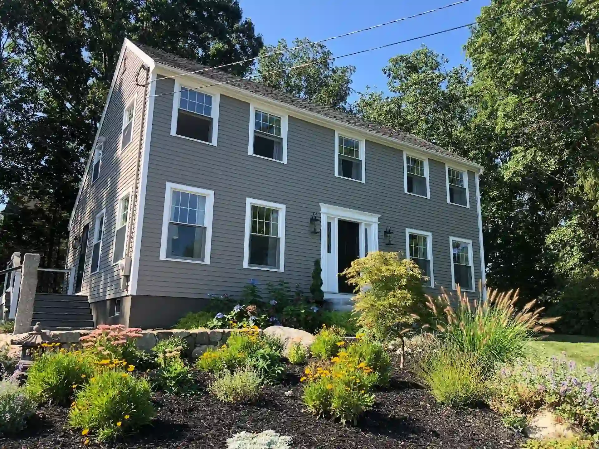 A multi-story gray house with white trim and several windows, surrounded by a landscaped garden with flowers and greenery, under a clear blue sky.