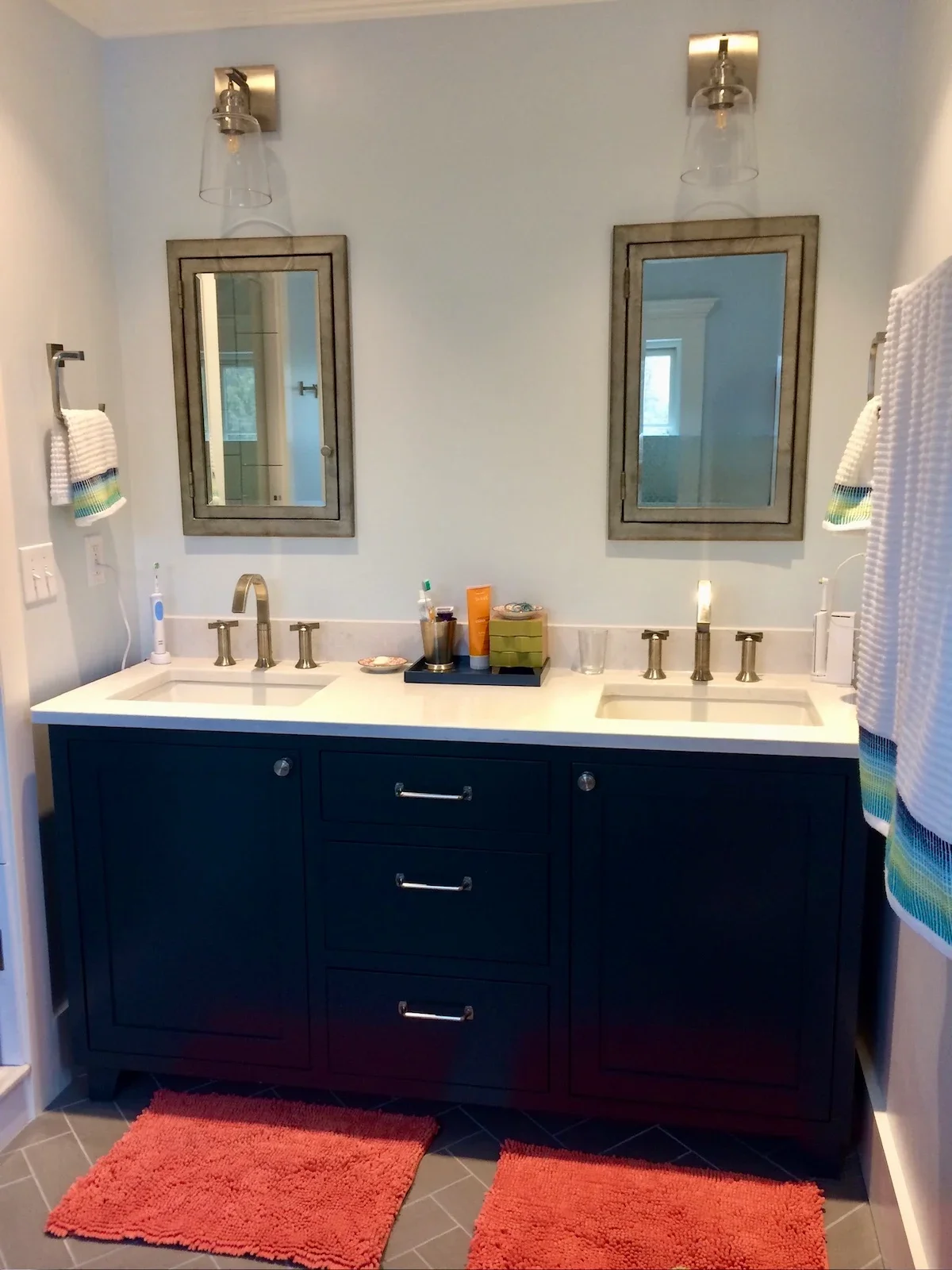 Modern bathroom vanity with two sinks, dark blue cabinet, white countertop, and two wall mirrors with light fixtures above each mirror. Items on the counter include an electric toothbrush, toiletries, a glass, and decorative objects. Two orange bath 